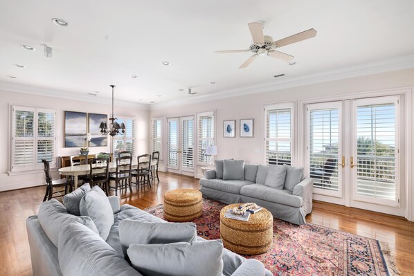 A spacious living room with a grey sofa, a dining table with chairs, a ceiling fan, and large windows with shutters.