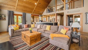 A spacious living room with a high ceiling, a large sofa set, a wooden coffee table, and a view of the kitchen and dining area. - Pet-Friendly Mountain View Retreat | Near Lake & Trails | Rec Center Access (Truckee)
