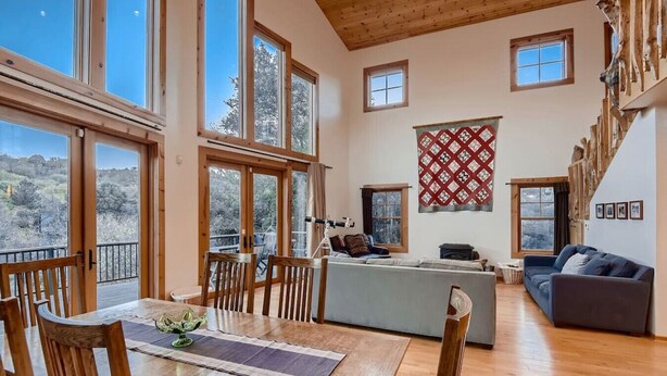 A spacious living area with a wooden dining table, chairs, a sofa, and a balcony with a view of trees. - Custom Built by the Littlehale Family (Julian)