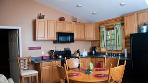 A compact kitchen with wooden cabinets, a microwave, a stove, and a dining area with a table and chairs. - Nicely Appointed Cabin in Scenic Setting. Family-friendly, Romantic Too!  (Arrington)