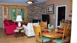 A cozy living room with a red sofa, a floral couch, a wooden dining table with chairs, a ceiling fan, and a stone wall. - Nicely Appointed Cabin in Scenic Setting. Family-friendly, Romantic Too!  (Arrington)