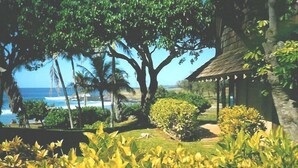 A tropical garden with a thatched-roof hut, lush greenery, and a view of the ocean. - Kaluako'i Resort Cottage 1A Beachfront Rental - Molokai, Hawaii (Maunaloa)
