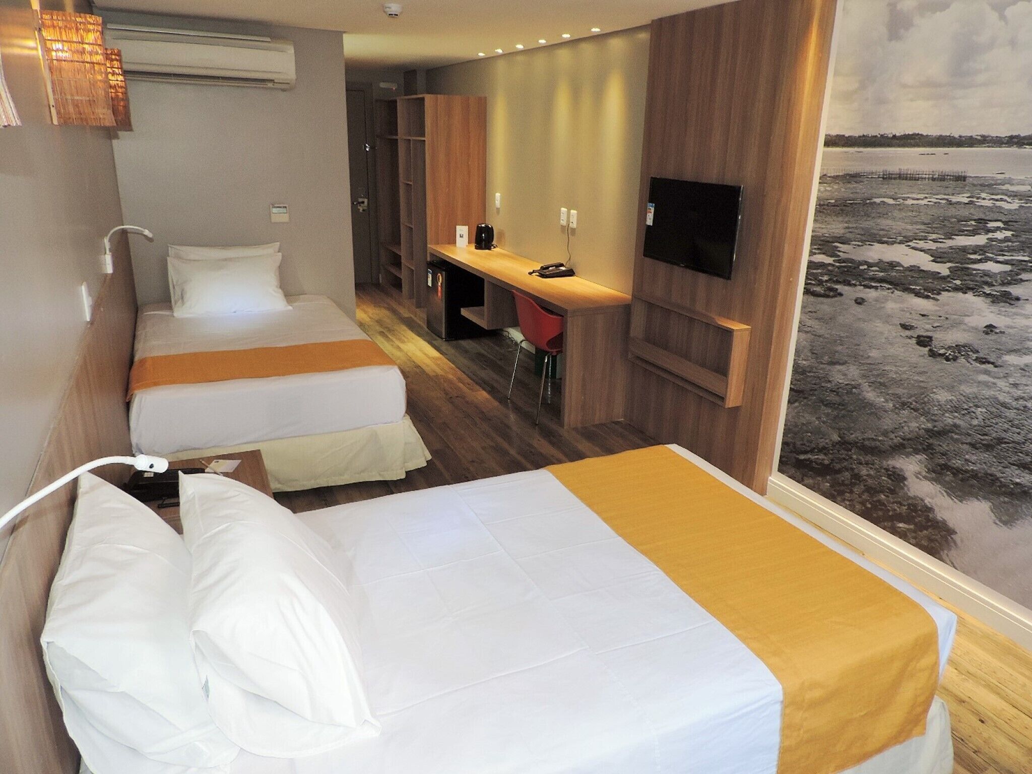 A hotel room with two beds, a desk, a TV, and a view of a beach.