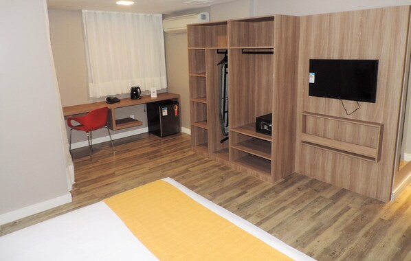 Habitación de hotel compacta con un armario de madera, un escritorio con silla, una kitchenette pequeña y un televisor montado en la pared.