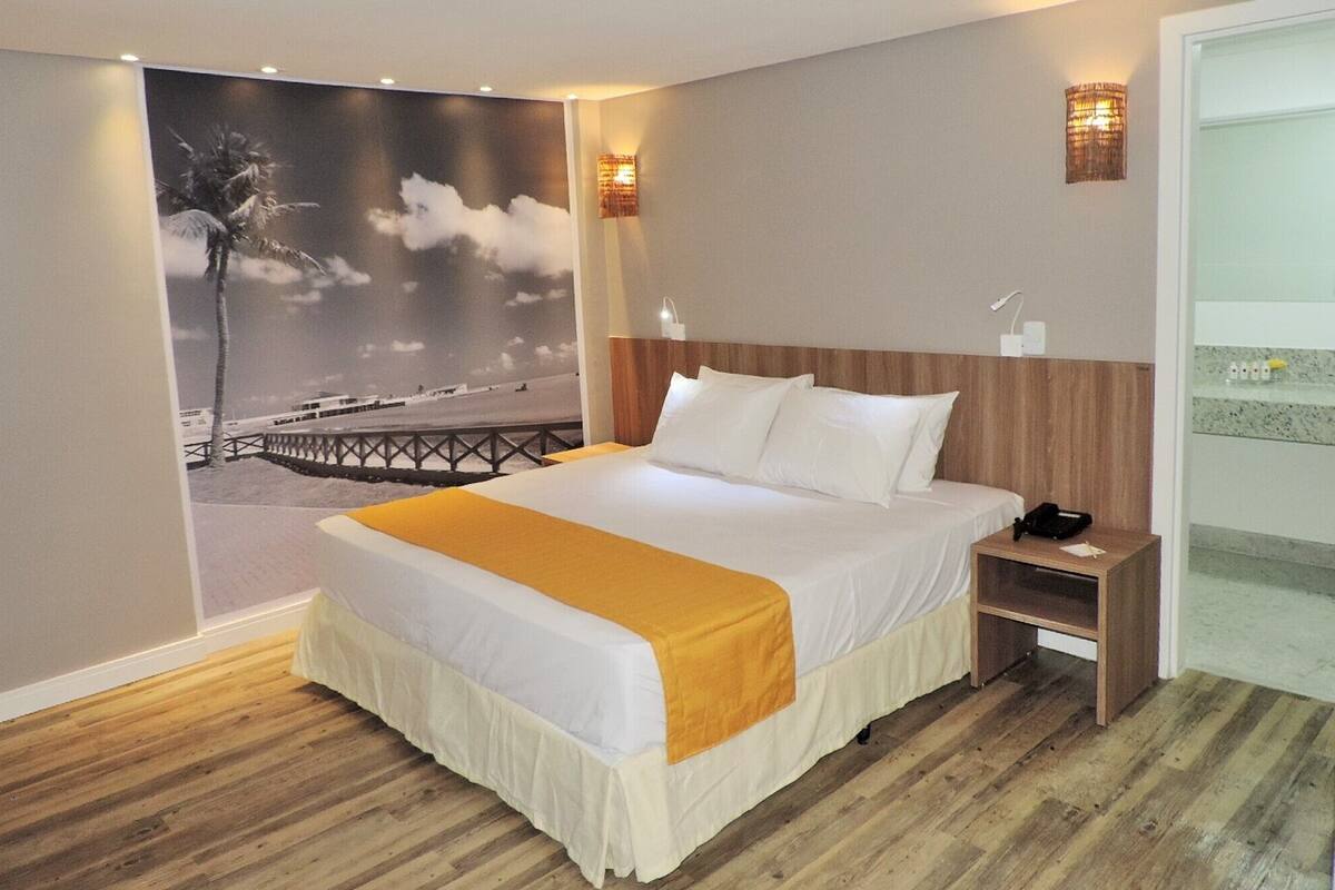 Une chambre d’hôtel avec un grand lit, une tête de lit en bois, deux tables de chevet, une petite table, une lampe fixée au mur et une affiche représentant une scène de plage.