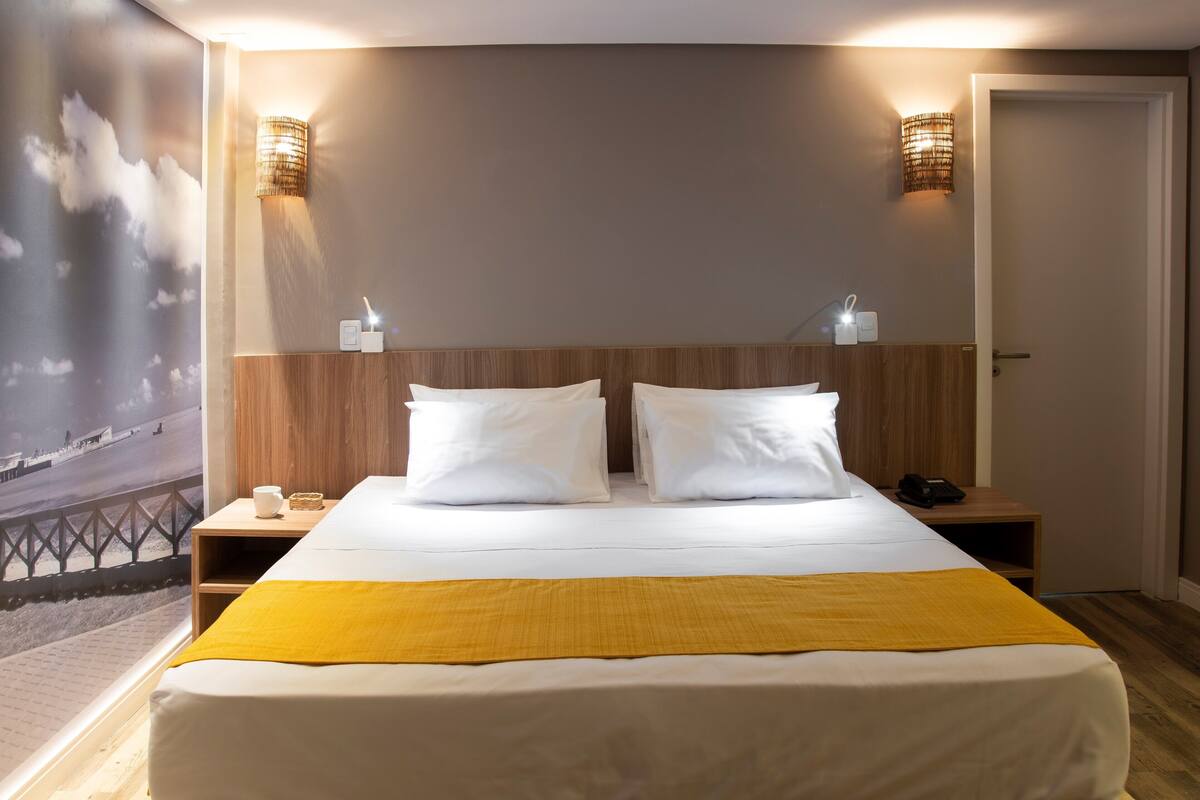 Un lit double avec du linge de lit blanc et un plaid jaune, deux tables de chevet avec des lampes et un luminaire fixé au mur.