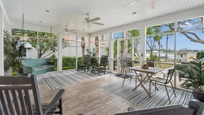 Um alpendre coberto com mesa e cadeiras, ventiladores de teto e grandes janelas com vista para uma área residencial.