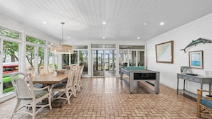 Uma sala de jantar ampla com mesa e cadeiras de madeira, mesa de bilhar e grandes janelas com vista para o exterior.