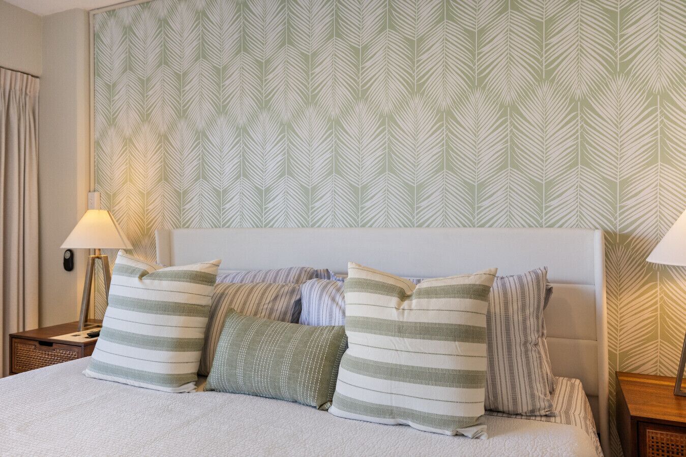 A neatly made bed with striped pillows and a headboard, a bedside lamp, and a patterned wallpaper.