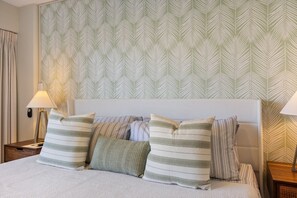 Una cama bien hecha con almohadas a rayas y un cabecero, una lámpara de noche y un papel tapiz estampado.