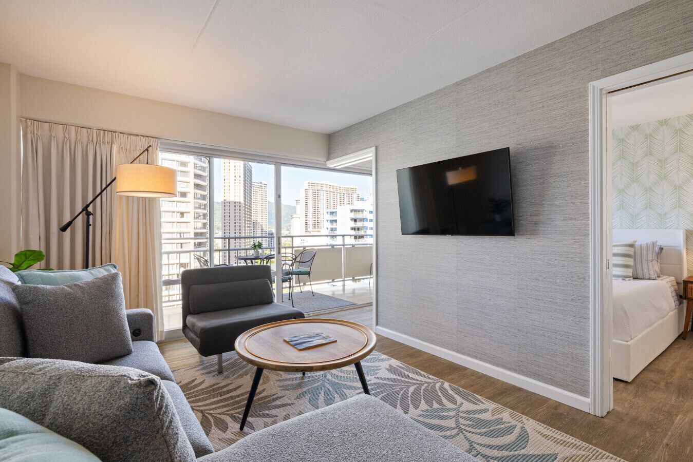 A modern hotel room with a balcony, a flat-screen TV, a sofa, a coffee table, and a bed.