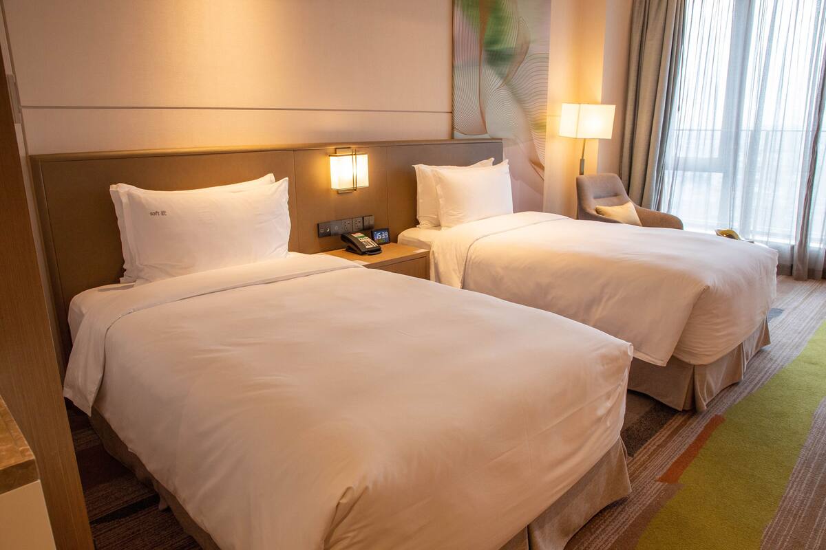 A hotel room with two single beds, a nightstand with a phone, a chair, and a window with curtains.