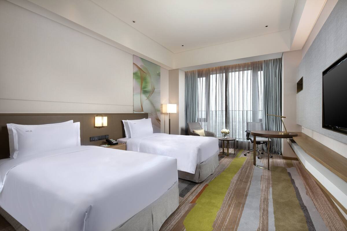 A hotel room with two beds, a desk, a chair, a TV, and a window with curtains.