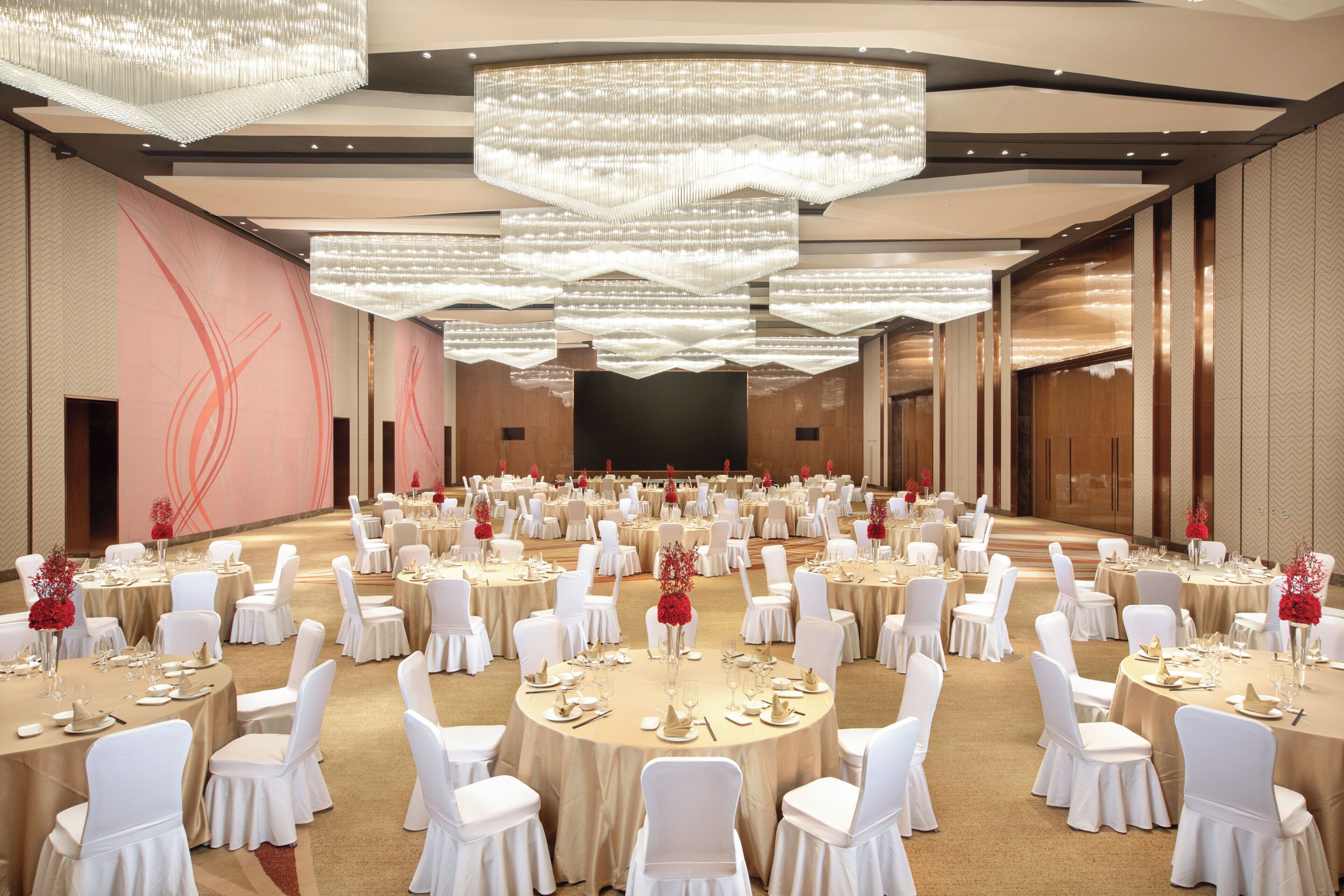 A banquet hall with round tables, white chairs, and chandeliers.