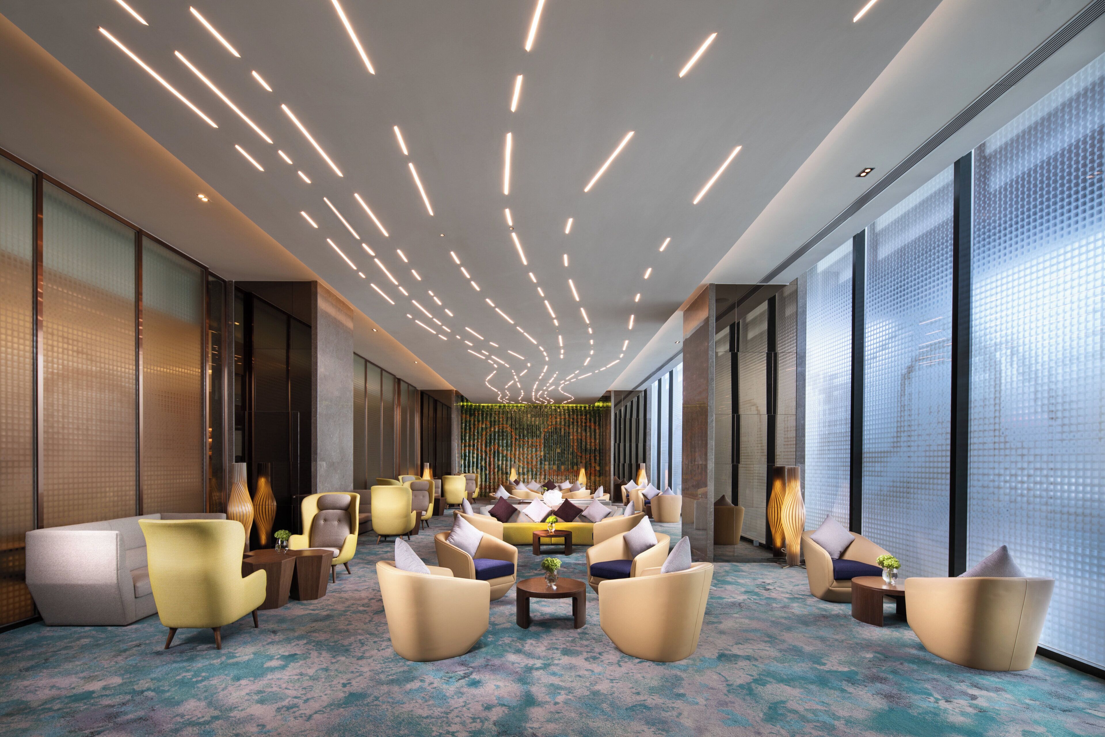 A modern lounge area with sofas and chairs arranged for relaxation, large windows providing natural light, and a patterned carpet.