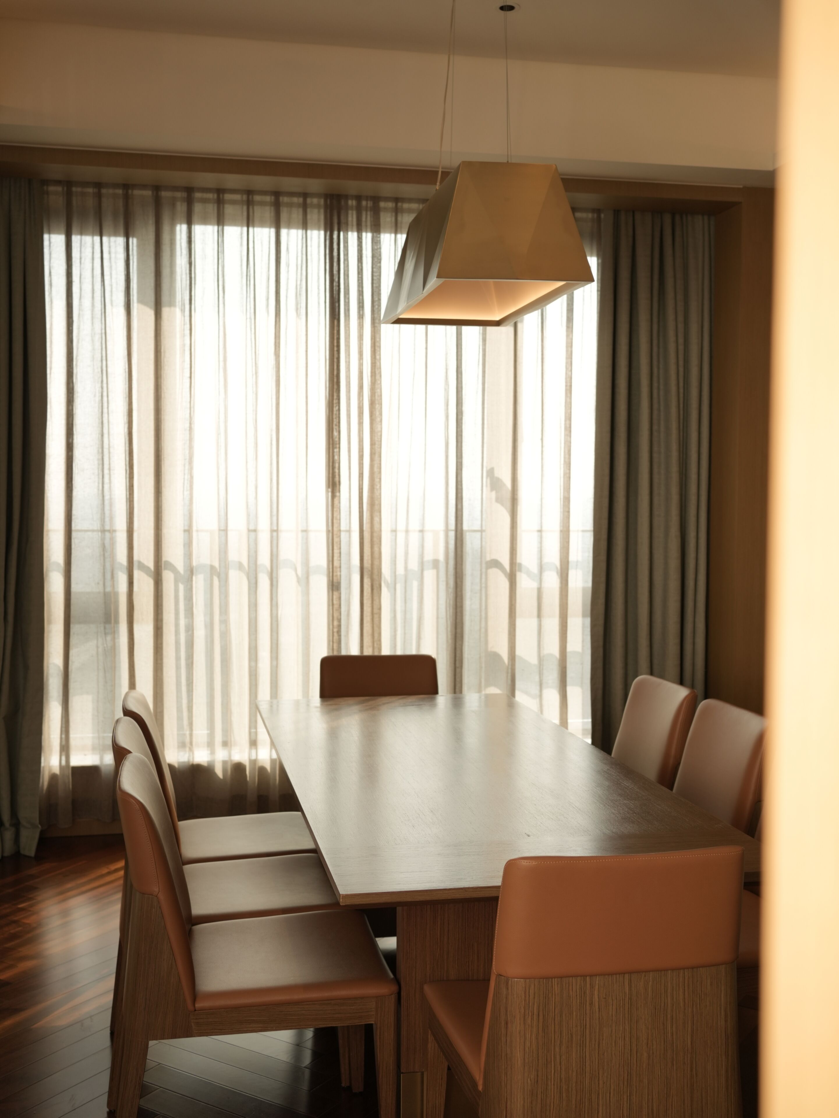 Uma área de refeições com mesa de madeira e seis cadeiras, janelas amplas com cortinas leves e um lustre.