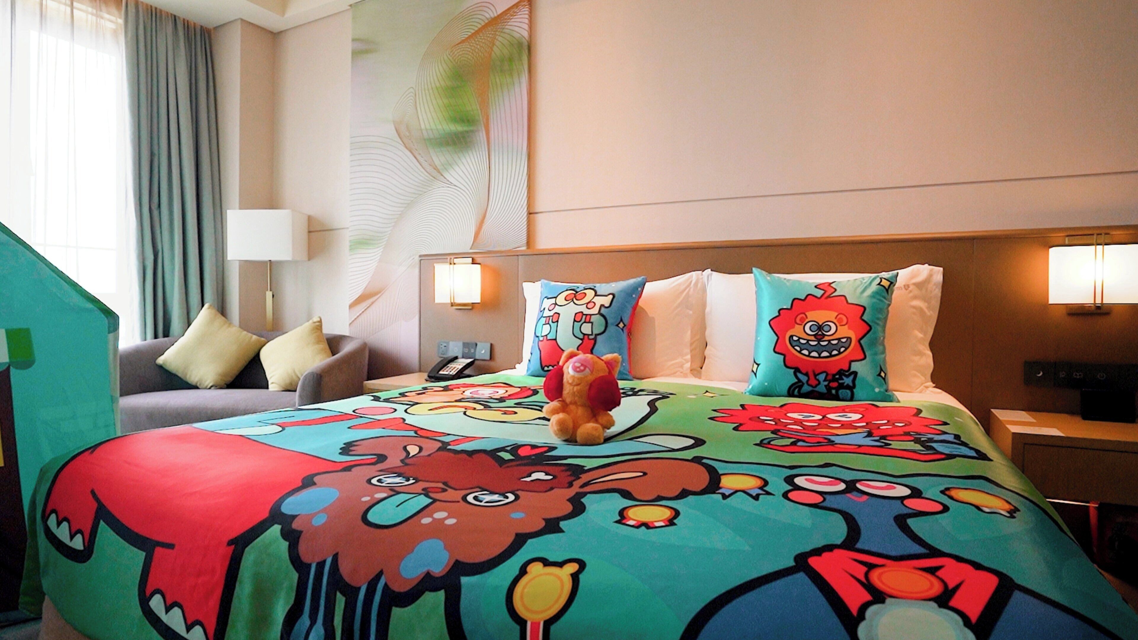 A hotel room with a colorful bedspread featuring cartoon characters, a sofa with cushions, a lamp, and a nightstand with a phone.