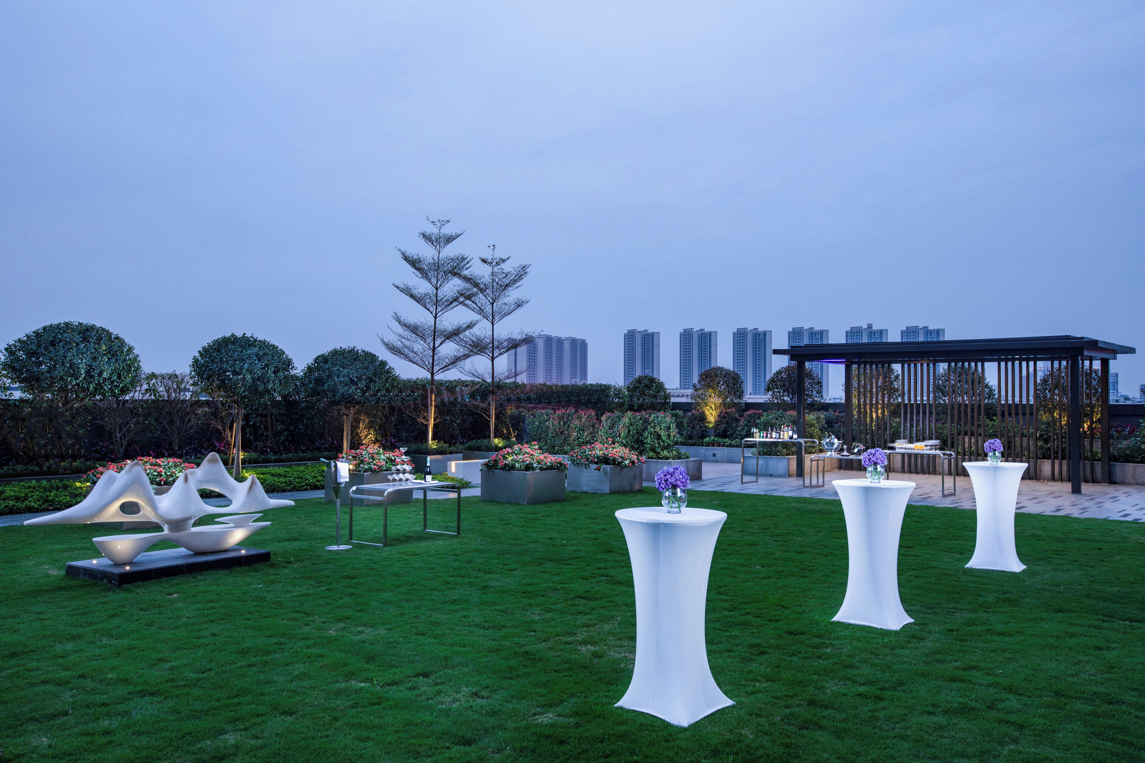 An outdoor dining area with white tables and chairs, surrounded by greenery and decorative elements.