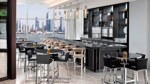 A modern restaurant with a city view, featuring a bar area with stools and a high ceiling with pendant lights. - Envue, Autograph Collection (Weehawken)