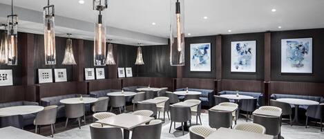 Ein modernes Restaurant mit runden Tischen, gepolsterten Stühlen, hängenden Pendelleuchten und abstrakter Kunst an den Wänden.