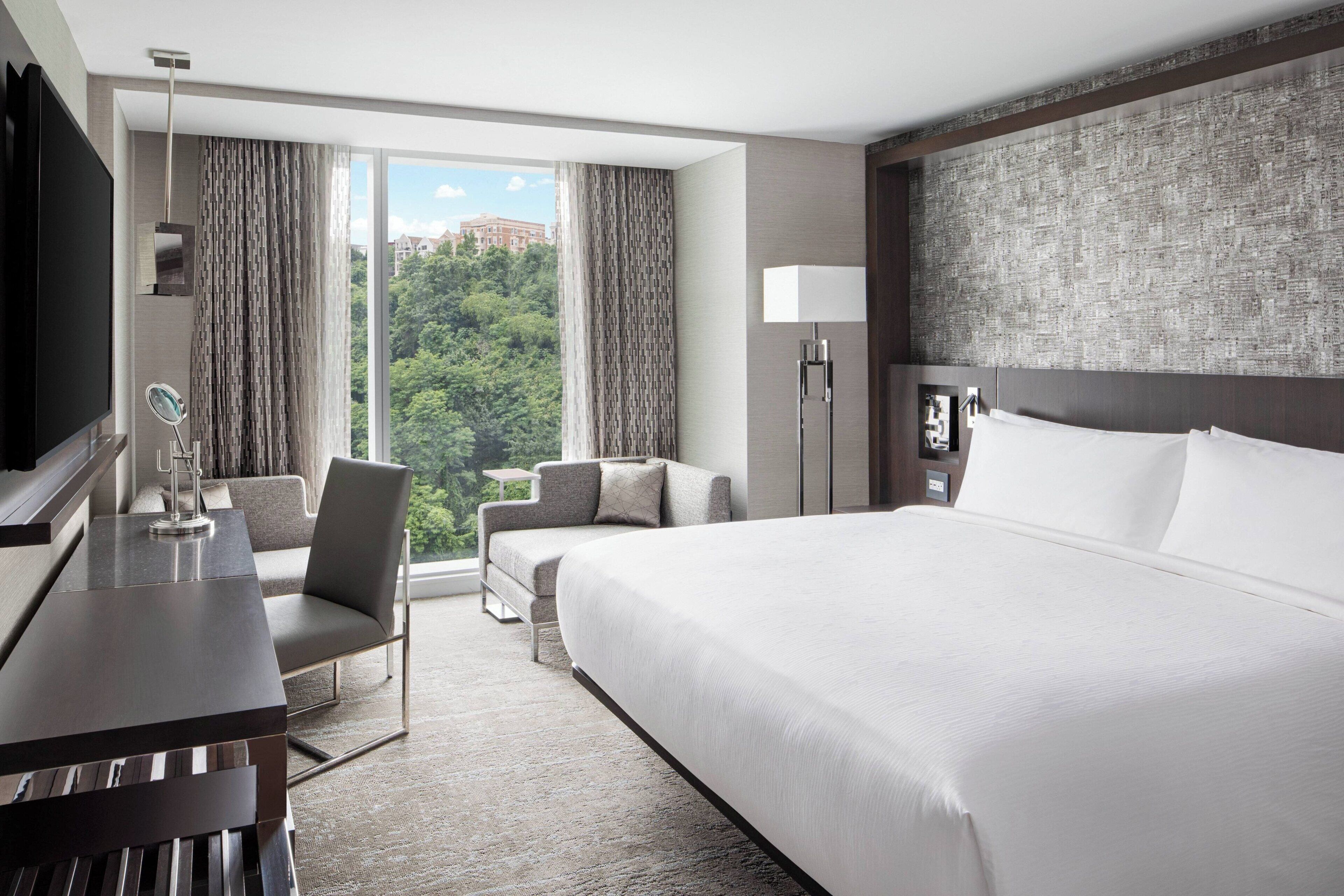 A hotel room with a large bed, a desk, a chair, a TV, and a view of greenery.