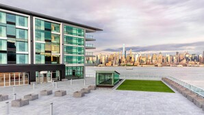 A modern building with large windows and a glass facade, situated near a waterfront with a city skyline in the background. - Envue, Autograph Collection (Weehawken)