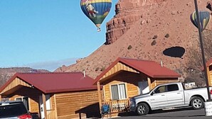 Une cabane en bois avec un toit rouge, un pick-up argenté garé à proximité, et un ciel bleu dégagé où flottent des montgolfières.