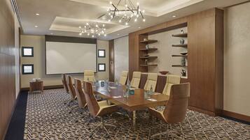 A conference room with a long table, chairs, a projector screen, and a decorative shelf with items.