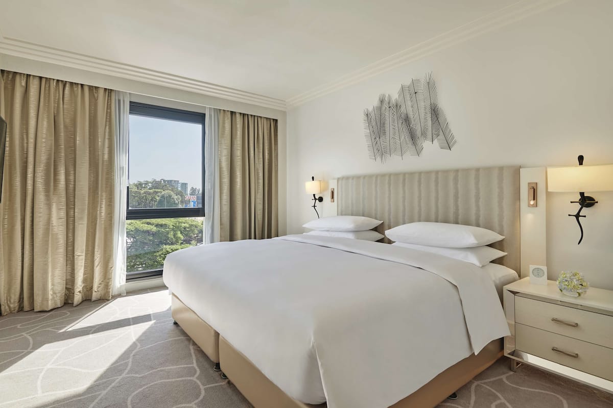 A hotel room with a large bed, a flat-screen TV mounted on the wall, a window with curtains, a wall-mounted lamp, and a small table with a vase.