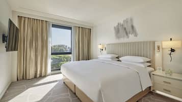 Ein Hotelzimmer mit einem großen Bett, einem flachbildfernseher an der Wand, einem Fenster mit Vorhängen, einer Wandlampe und einem kleinen Tisch mit einer Vase.