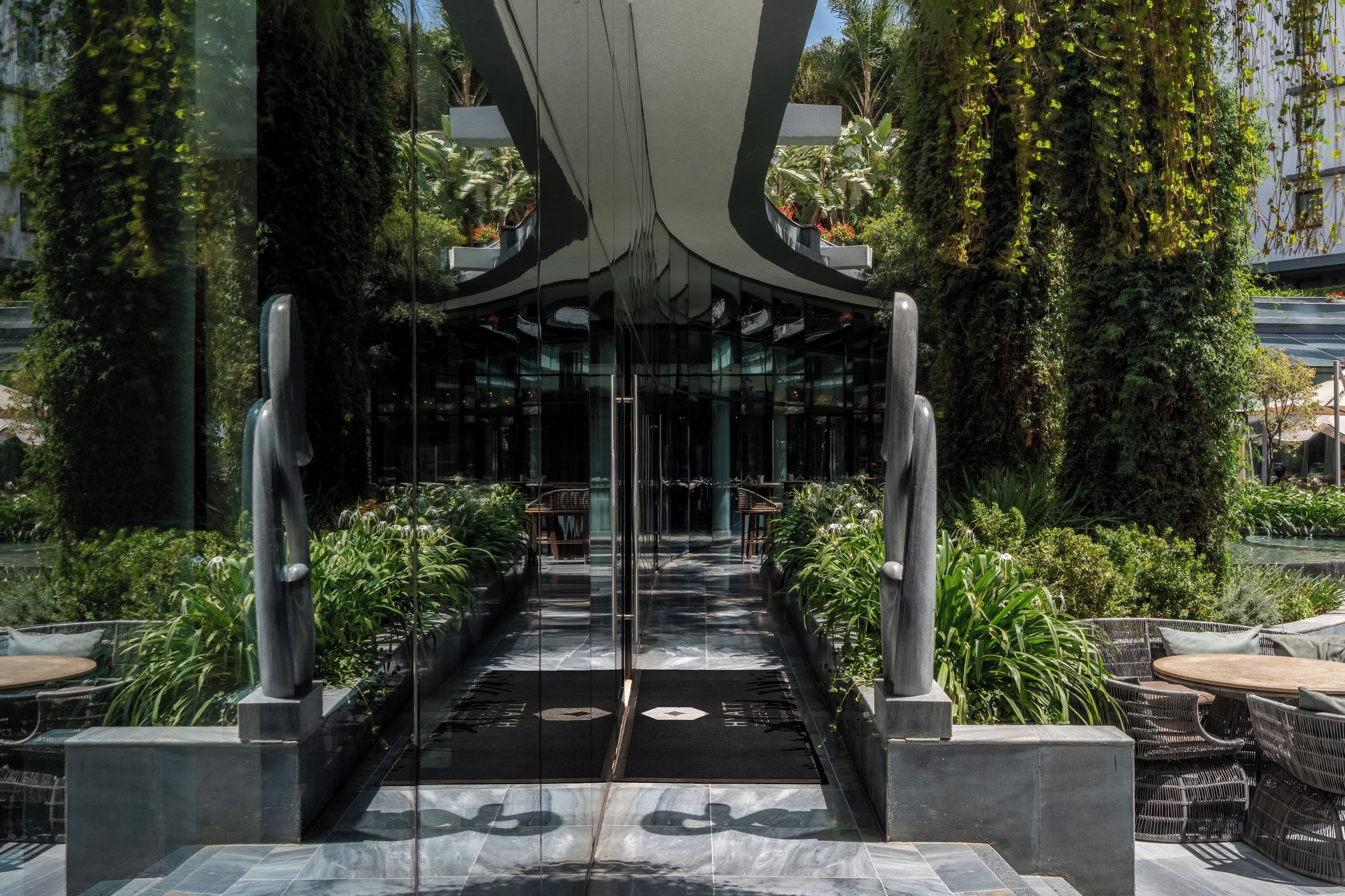 A modern building entrance with a glass facade, a central pathway leading to the entrance, and outdoor seating areas on either side.