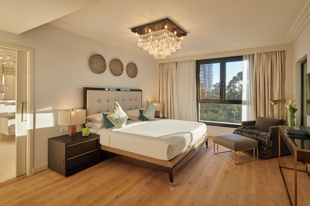A modern hotel room with a large bed, a chandelier, a desk with a chair, and a large window offering a view.