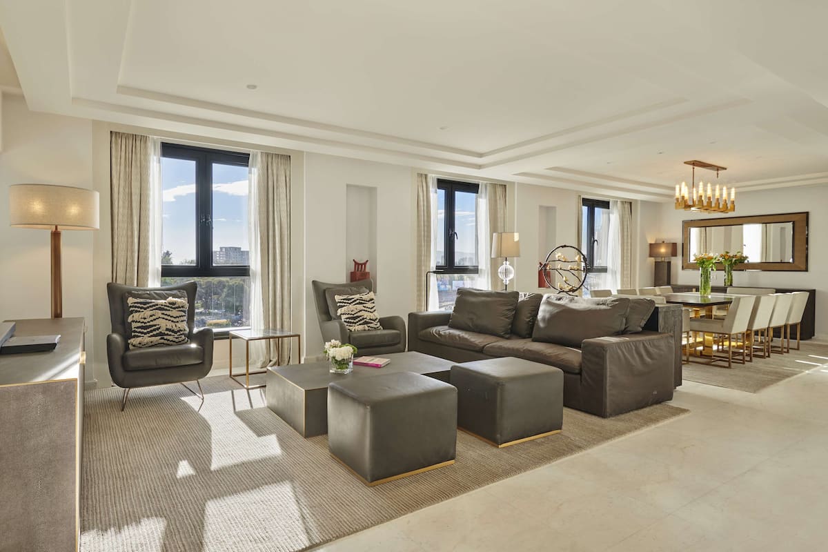 A modern living room with a flat-screen TV, comfortable seating, a dining area, and large windows offering a city view.