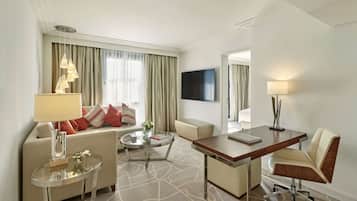 A modern hotel room with a sofa, a glass coffee table, a desk with a chair, a flat-screen TV mounted on the wall, and a lamp on the desk.