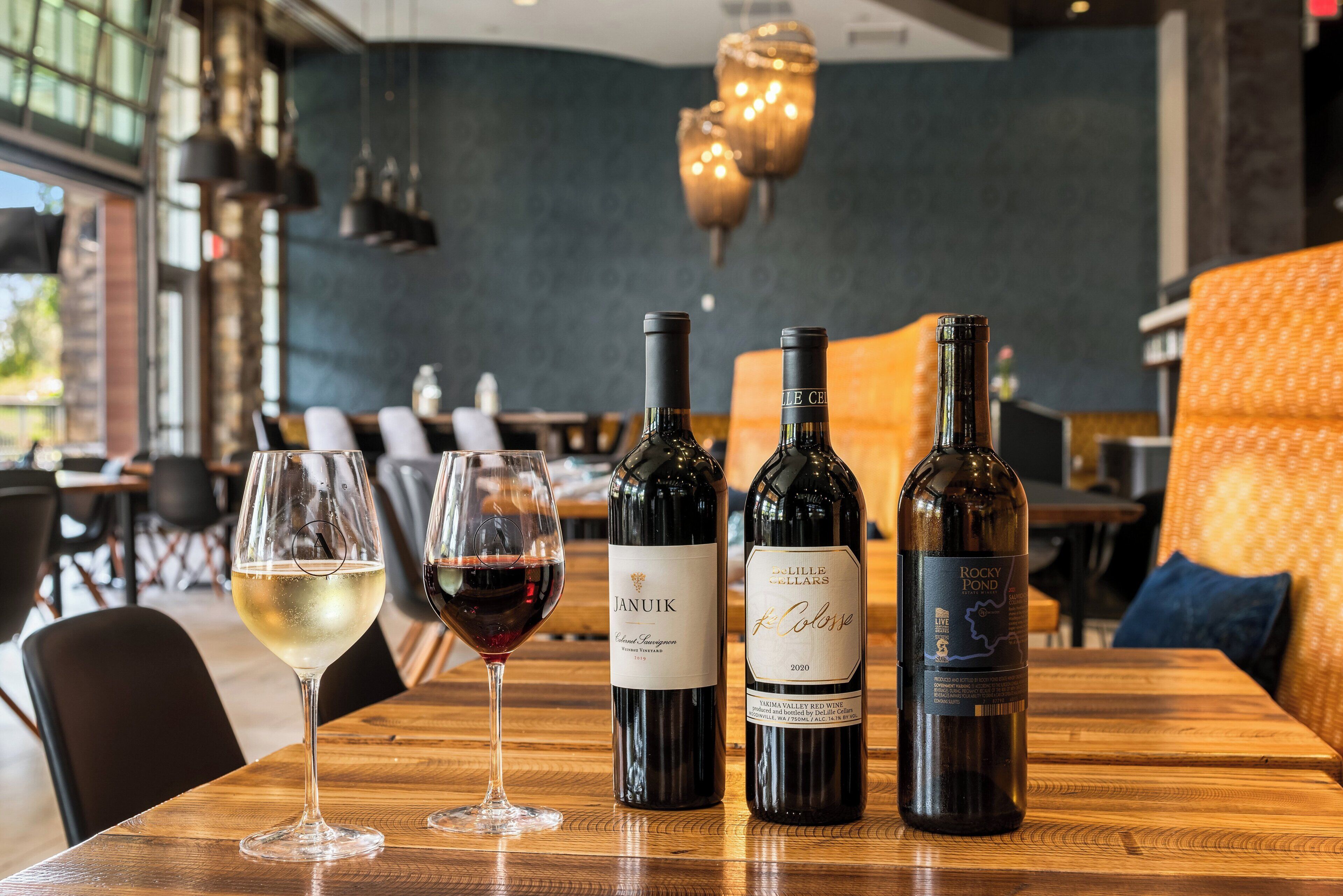 Una mesa de madera con dos botellas de vino y dos copas. El ambiente parece ser un restaurante o bar de estilo moderno, con paredes oscuras y muebles de madera.