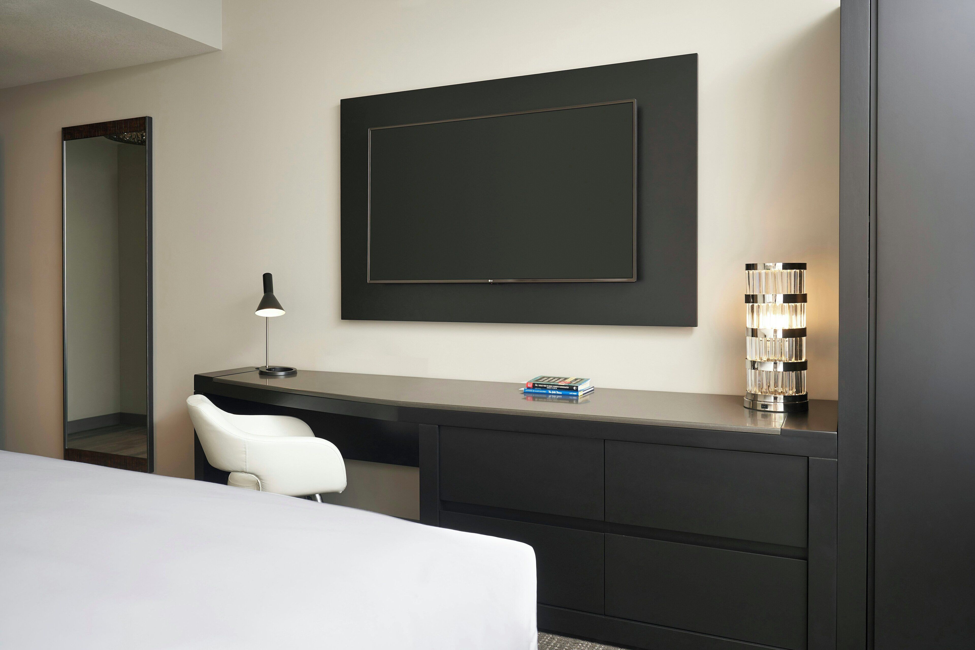 A modern hotel room with a large bed, a desk with a chair, a large mirror, a framed artwork, and a decorative lamp.