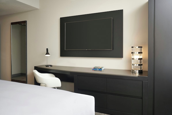 Una habitación de hotel moderna con una cama grande, un escritorio con una silla, un espejo amplio, una obra de arte enmarcada y una lámpara decorativa.