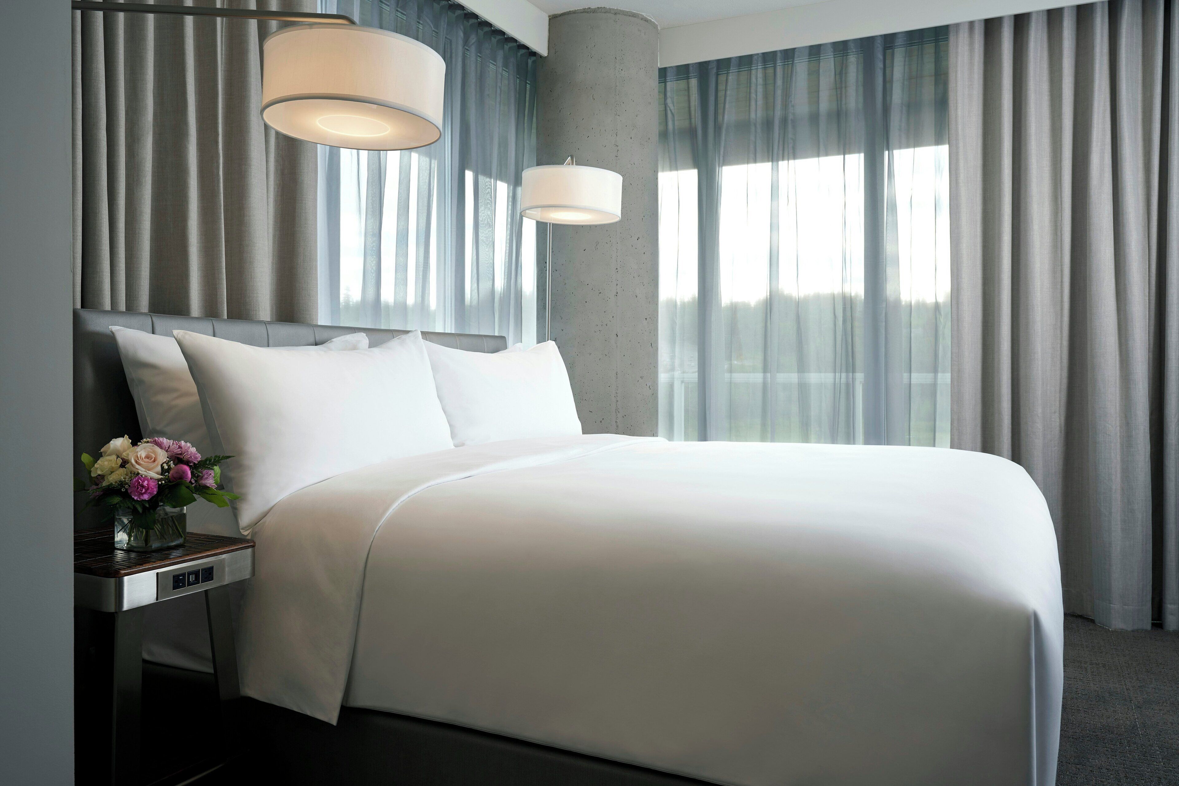 A modern hotel room with a large bed, a nightstand with a vase of flowers, and a large window with curtains.