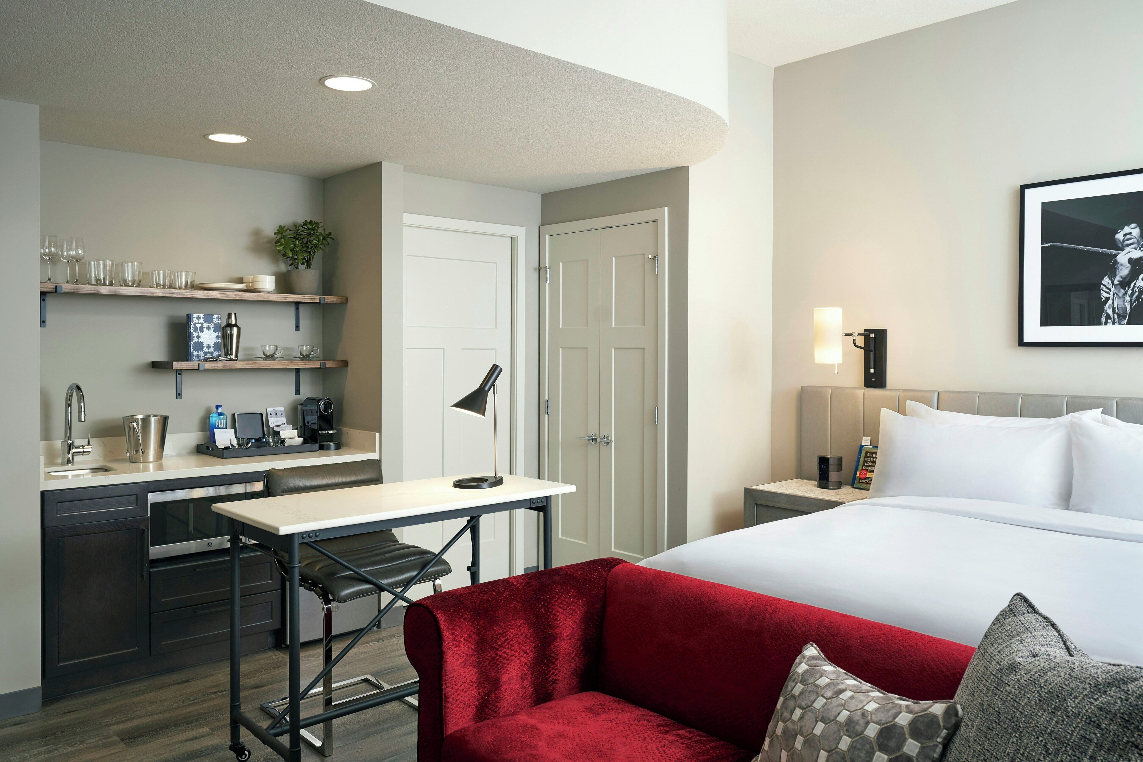 Une chambre d’hôtel comprenant un lit, un bureau avec une chaise, un canapé rouge, une petite table, une machine à café et une étagère avec des verres et une plante.
