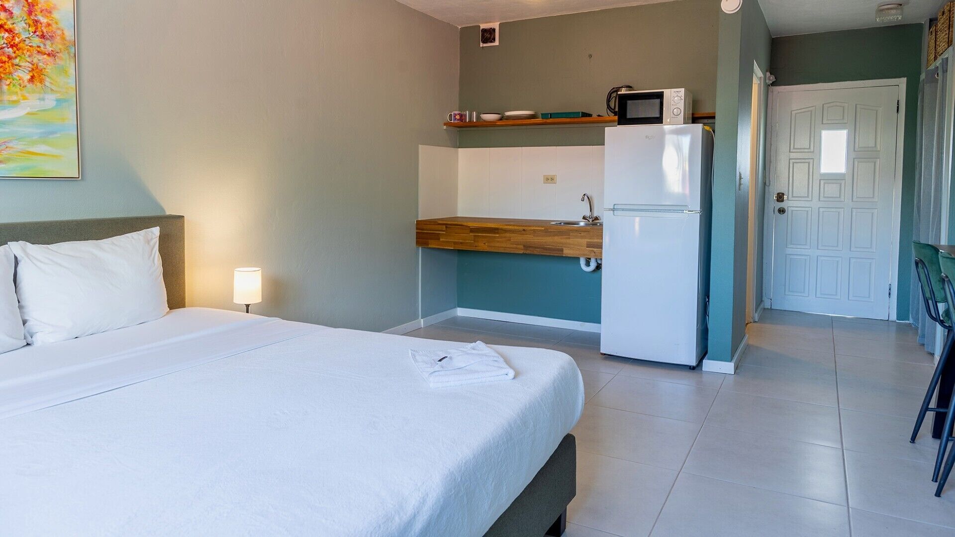 A hotel room with a bed, a small kitchenette with a microwave and refrigerator, a sink, and a door leading to another room.