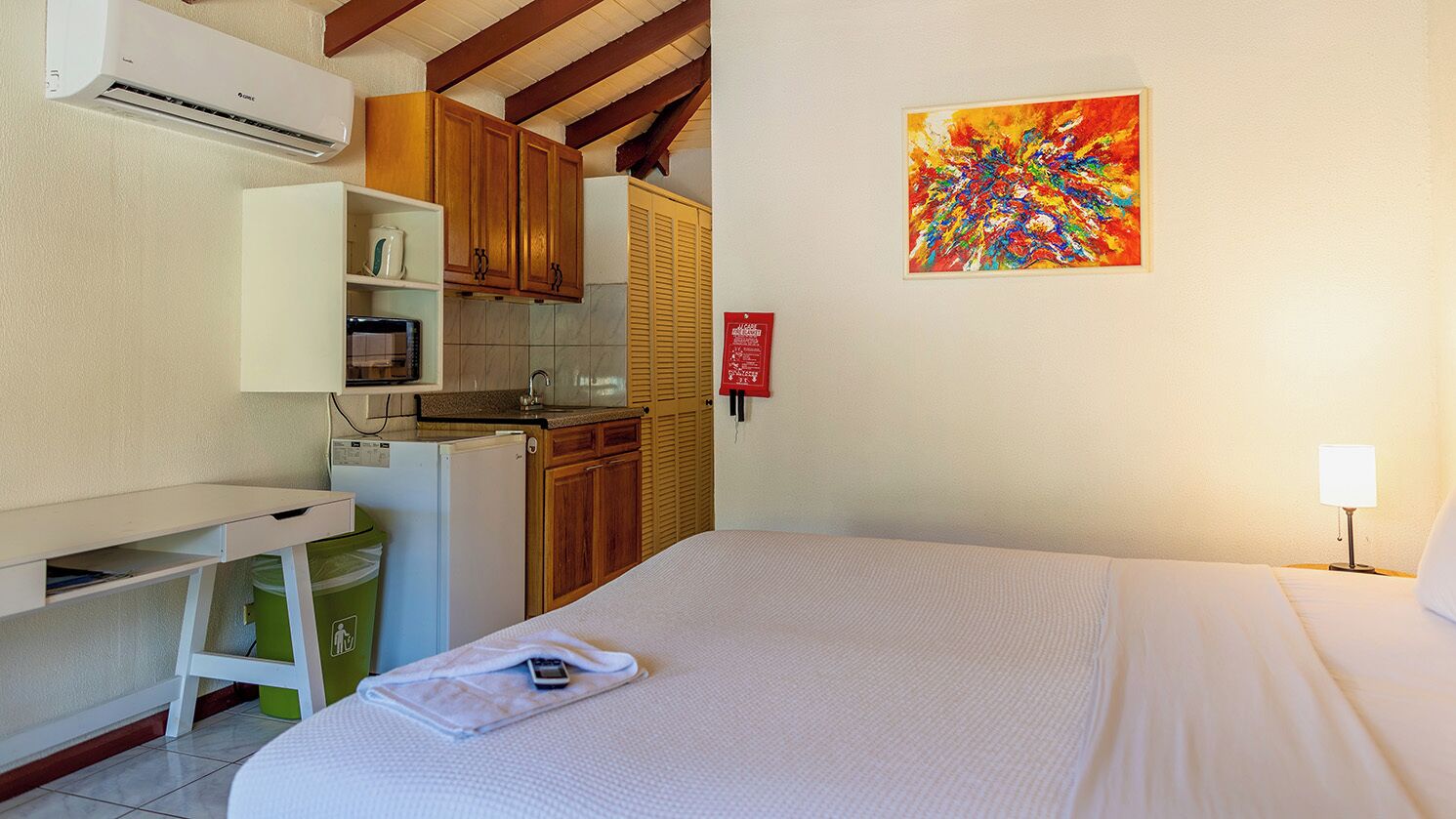 A hotel room with a bed, a desk, a kitchenette, and a colorful abstract painting on the wall.