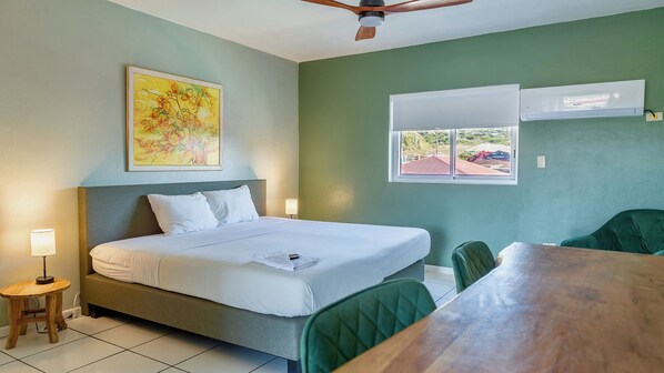 Habitación de hotel con cama, escritorio, silla, ventilador de techo, ventana con aire acondicionado y un cuadro en la pared.