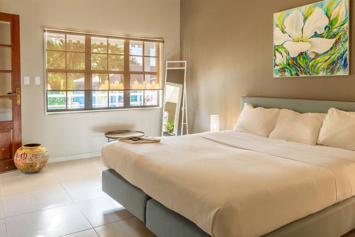 A bedroom with a large bed, a window with a view, a painting on the wall, and a wooden door.