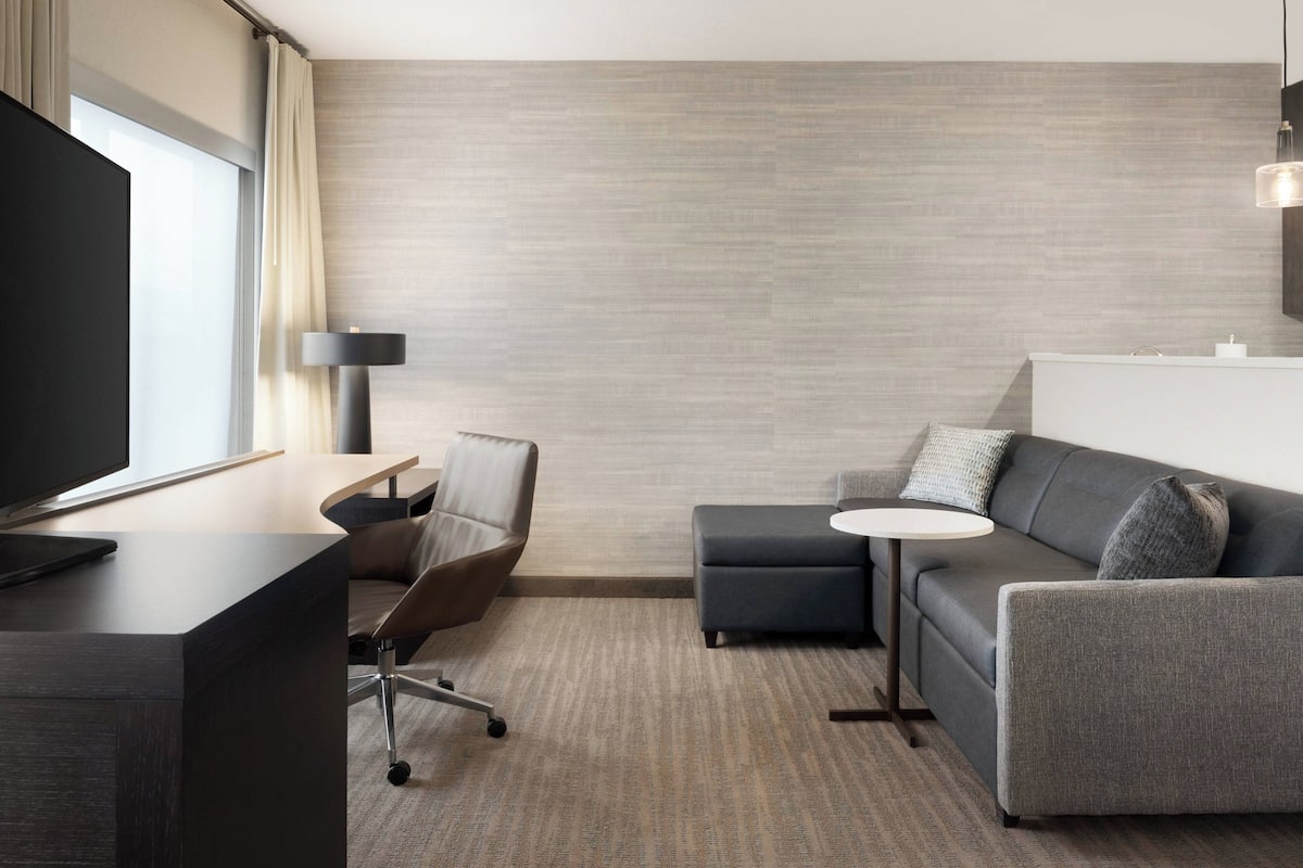 A modern hotel room with a flat-screen TV, a desk with a chair, a grey sofa, a small round table, and a lamp.