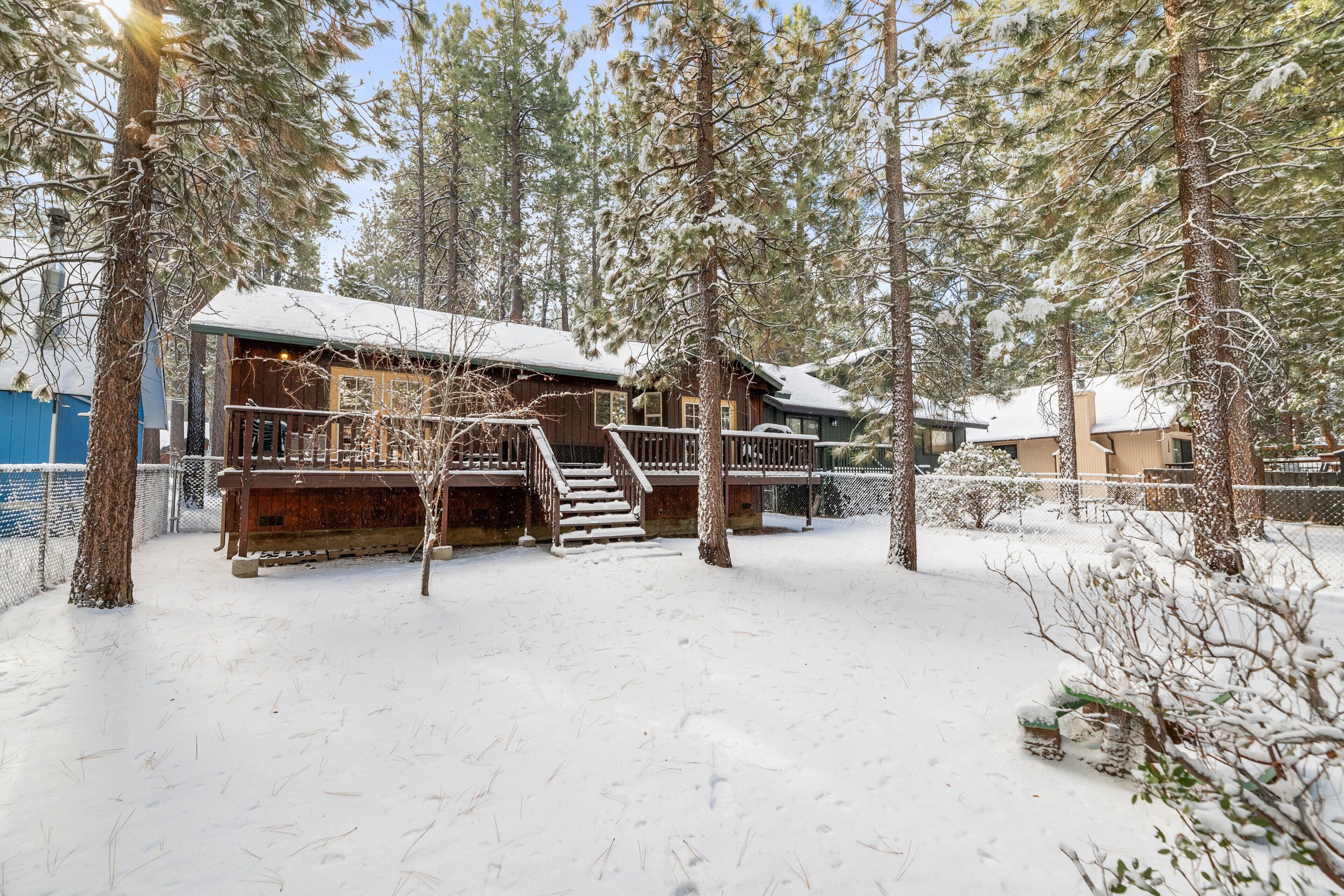 Een houten chalet met een overdekt terras en een trap naar een veranda, omringd door besneeuwde grond en bomen.