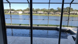 A view through a window of a swimming pool area with a clear view of a lake and houses in the background.