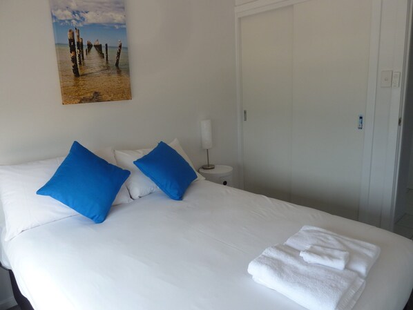 A hotel room with a bed, white bedding, two blue pillows, a white bedside table with a lamp, and a door.
