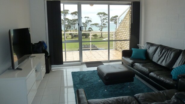 A living room with a black leather sofa, a flat-screen TV, and a large sliding glass door leading to a patio with a view of trees and a building.