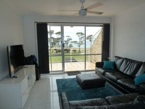 A living room with a black leather sofa, a flat-screen TV, and a large sliding glass door leading to a patio with a view of trees and a building. - Conveniently located on Bridport's Main St is this neat 3... (Bridport)