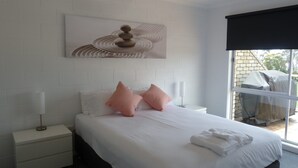 A hotel room with a bed, white nightstands, a ceiling fan, a window with a view of the outside, and a wall-mounted artwork.